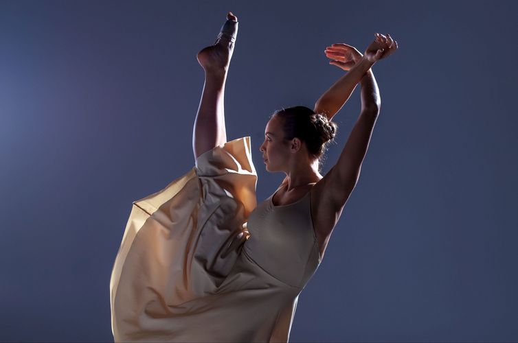 Balletttänzerin vor neutralem schlichten Hintergrund, ein Bein und die Arme sind im Tanz nach oben gestreckt.